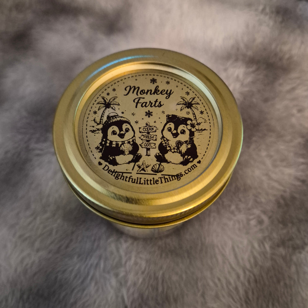 A round gold metal tin with a lid printed with a black penguin illustration and the text 'Monkey Farts' and 'DelightfulLittleThings.com', photographed from above on a gray faux-fur surface.