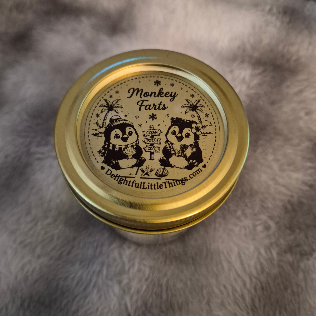 A round gold metal tin with a lid printed with a black penguin illustration and the text 'Monkey Farts' and 'DelightfulLittleThings.com', photographed from above on a gray faux-fur surface.