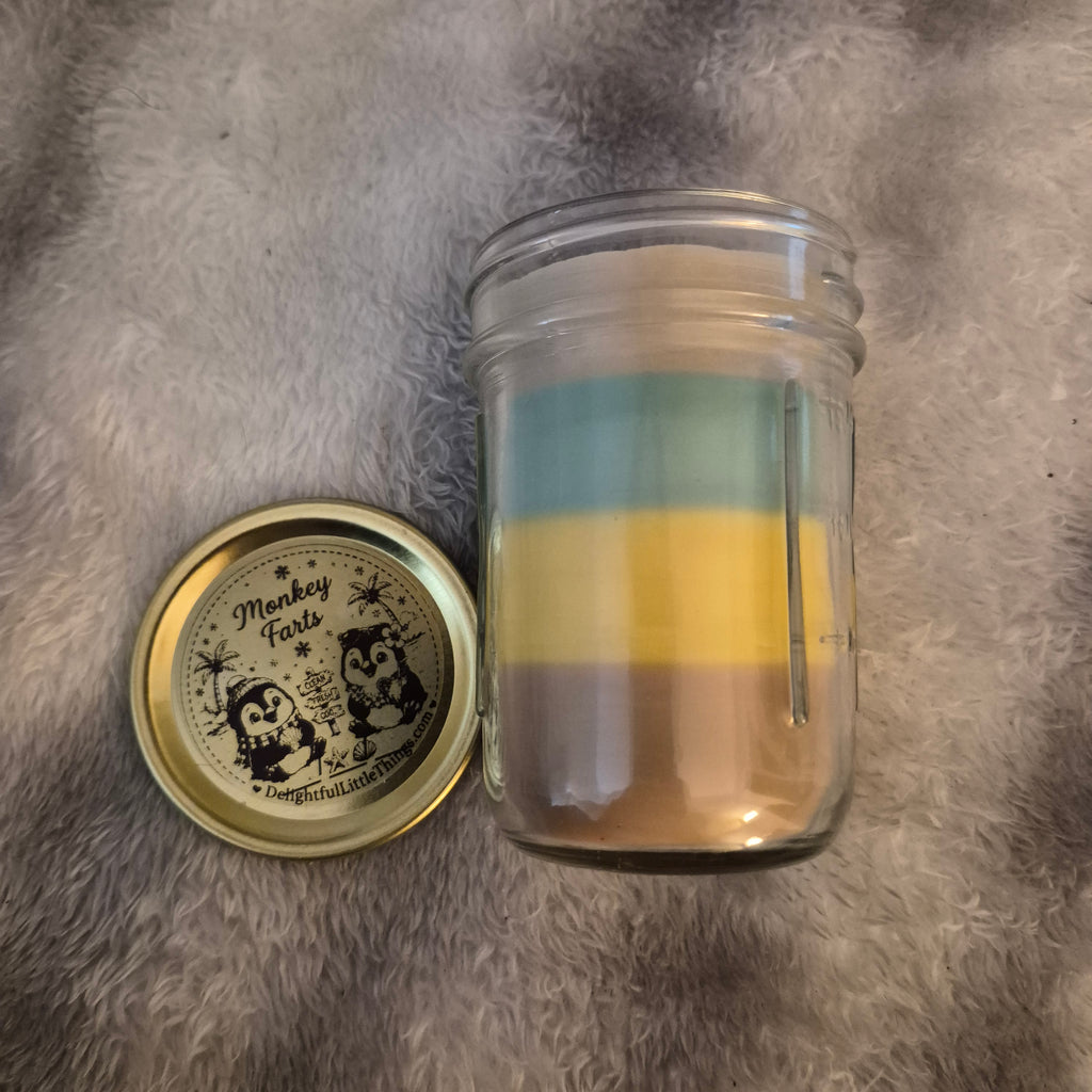 "9oz Monkey Farts scented handmade candle in glass jar with black lid, light green layered wax and Delightful Little Things Butt Naked label with penguin beach scene, DelightfulLittleThings.com"